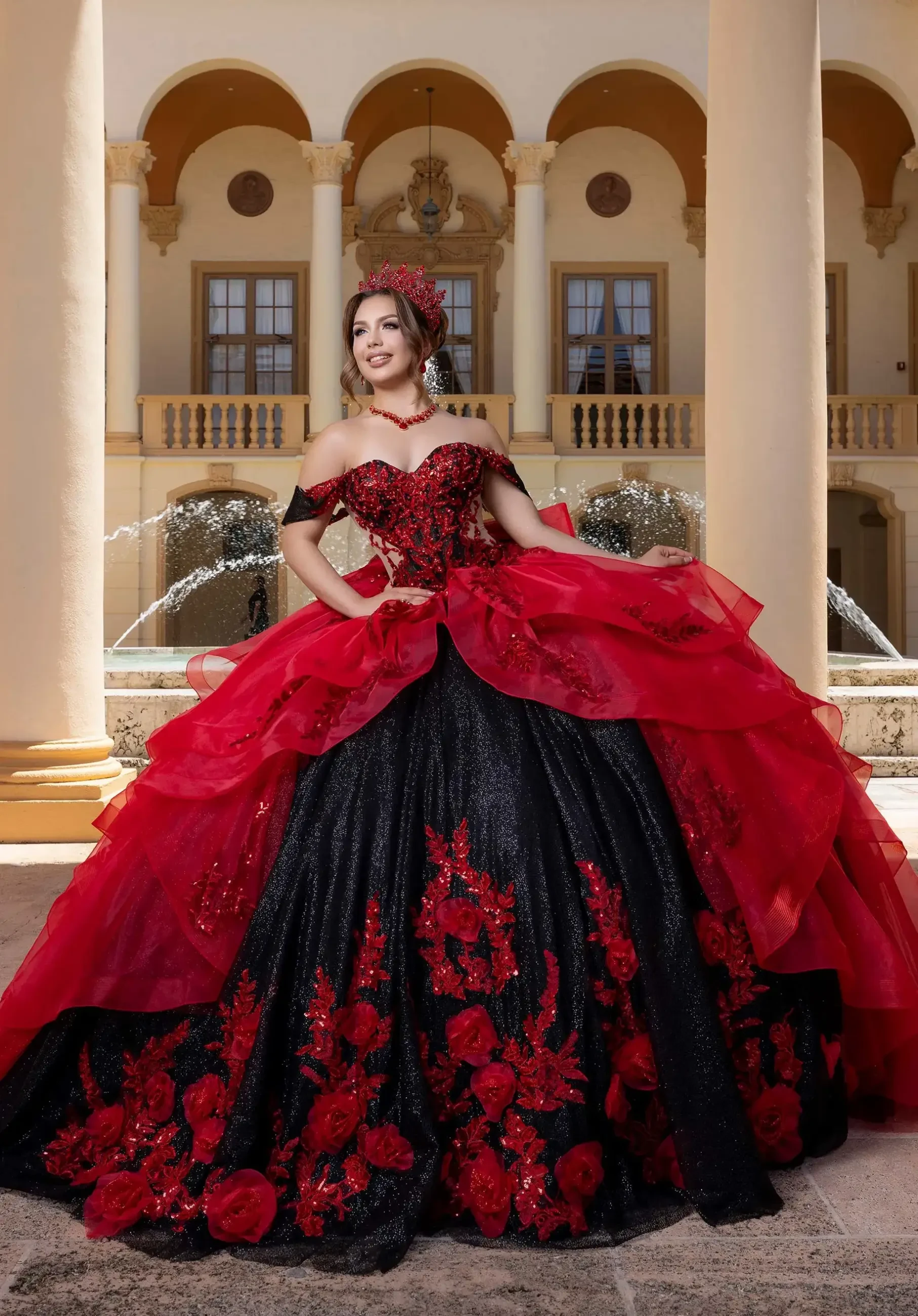 A woman in a black and red ball gown adorned with floral designs stands confidently in front of a grand building with columns and fountains.