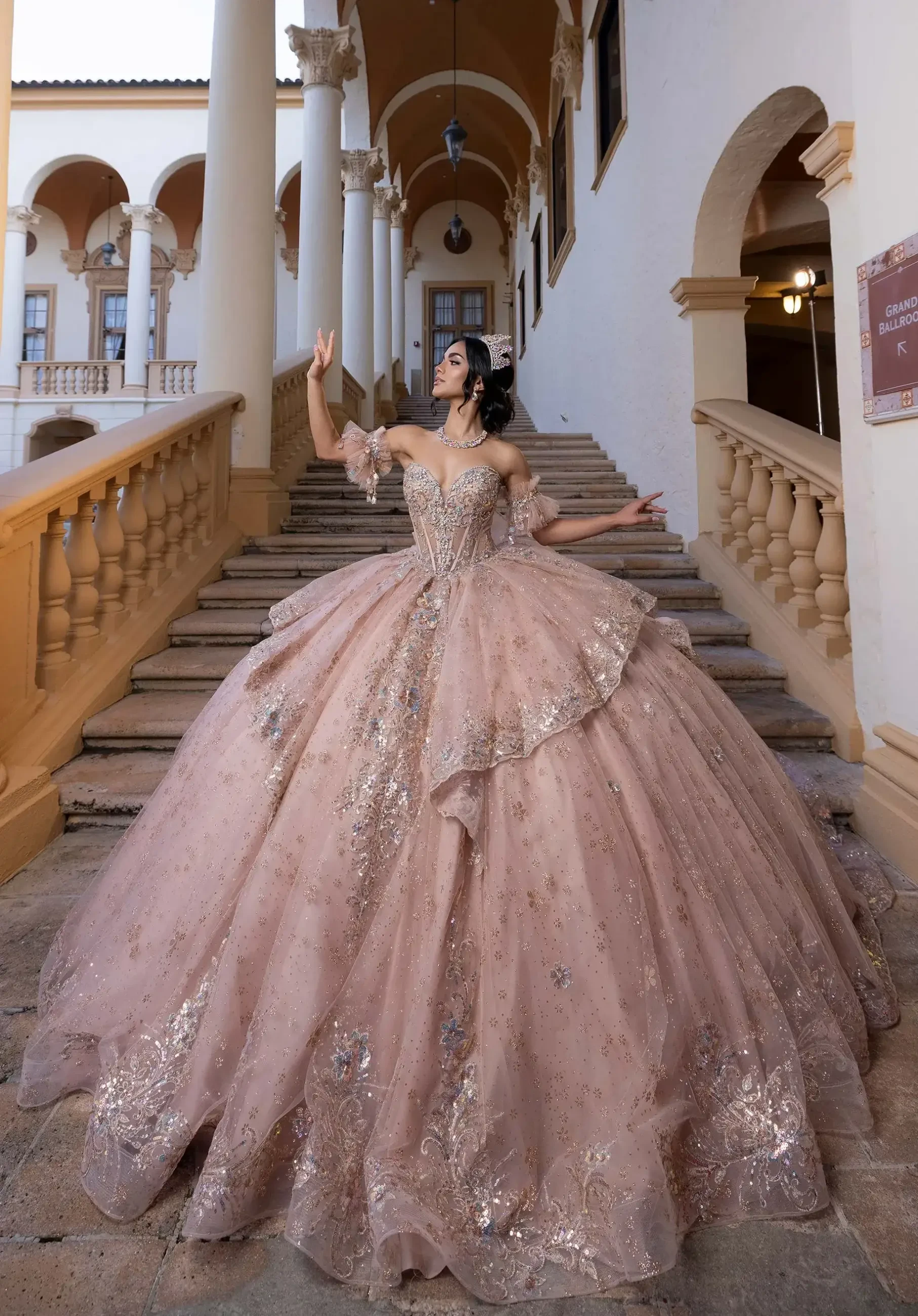 A woman in a large, elegant gown stands on a staircase, posing gracefully. The gown is pink with intricate designs and embellishments. The setting features an ornate architectural background with columns and decorative details.