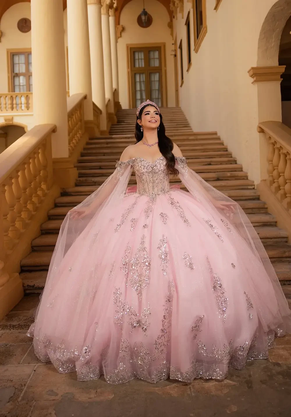 A young woman in a pink gown adorned with silver embellishments, standing on a staircase in a grand hall.