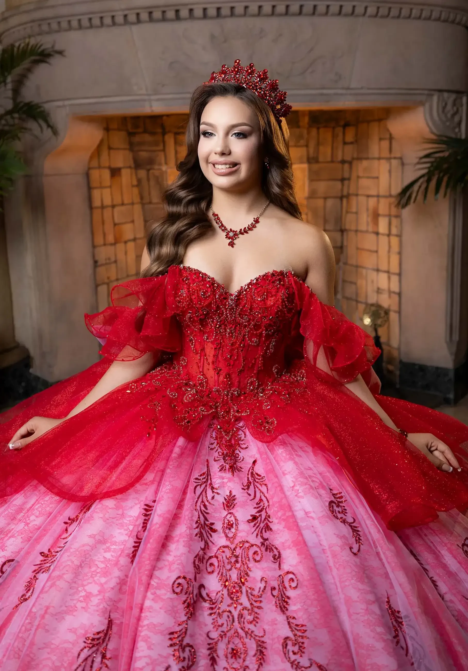A young woman wearing an elaborate red and pink gown adorned with sequins and lace, standing in front of a fireplace. She has a tiara on her head and matching jewelry, with long hair styled in loose waves.