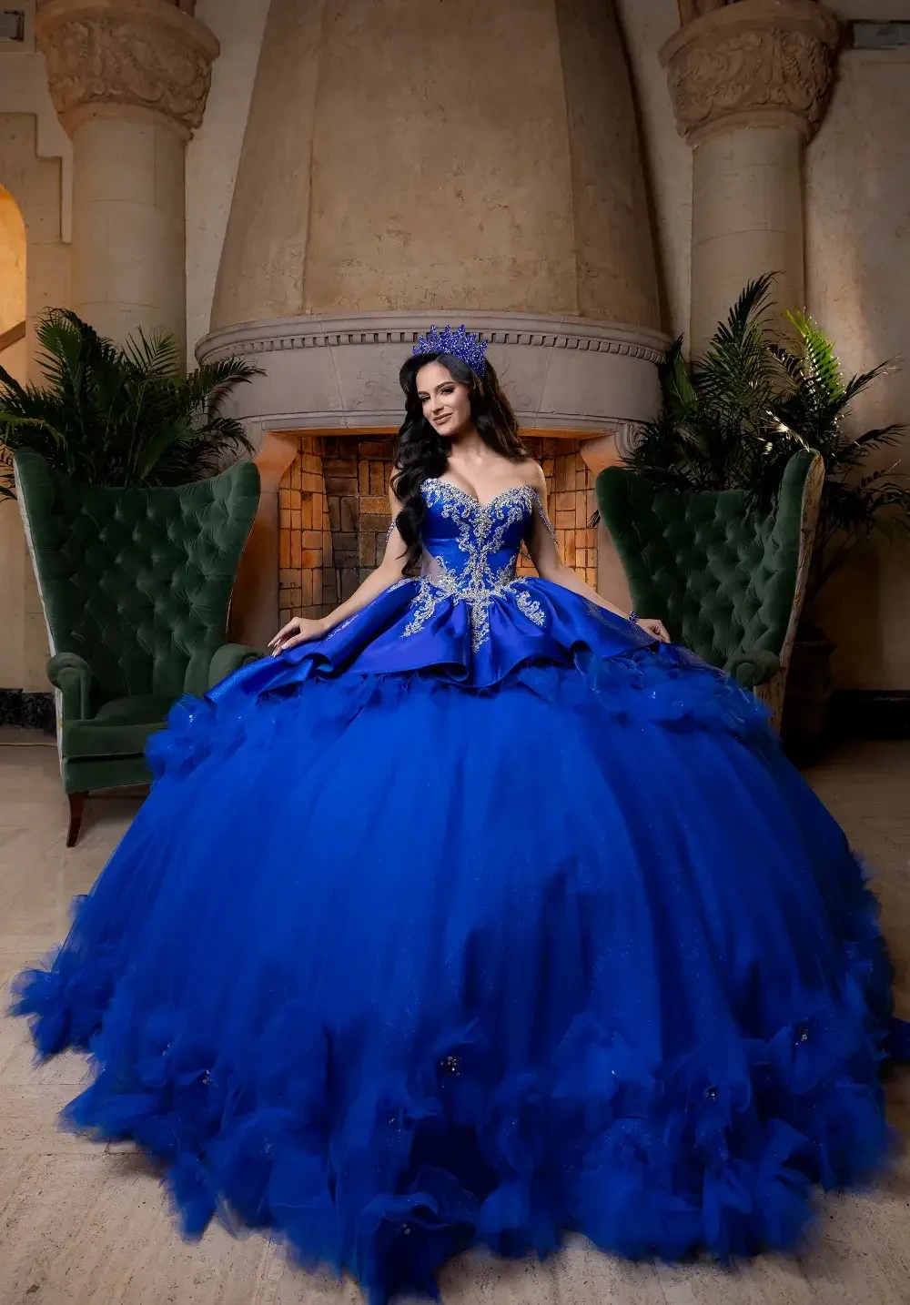 A woman in a stunning blue ball gown stands in a grand room with large green chairs and decorative plants.