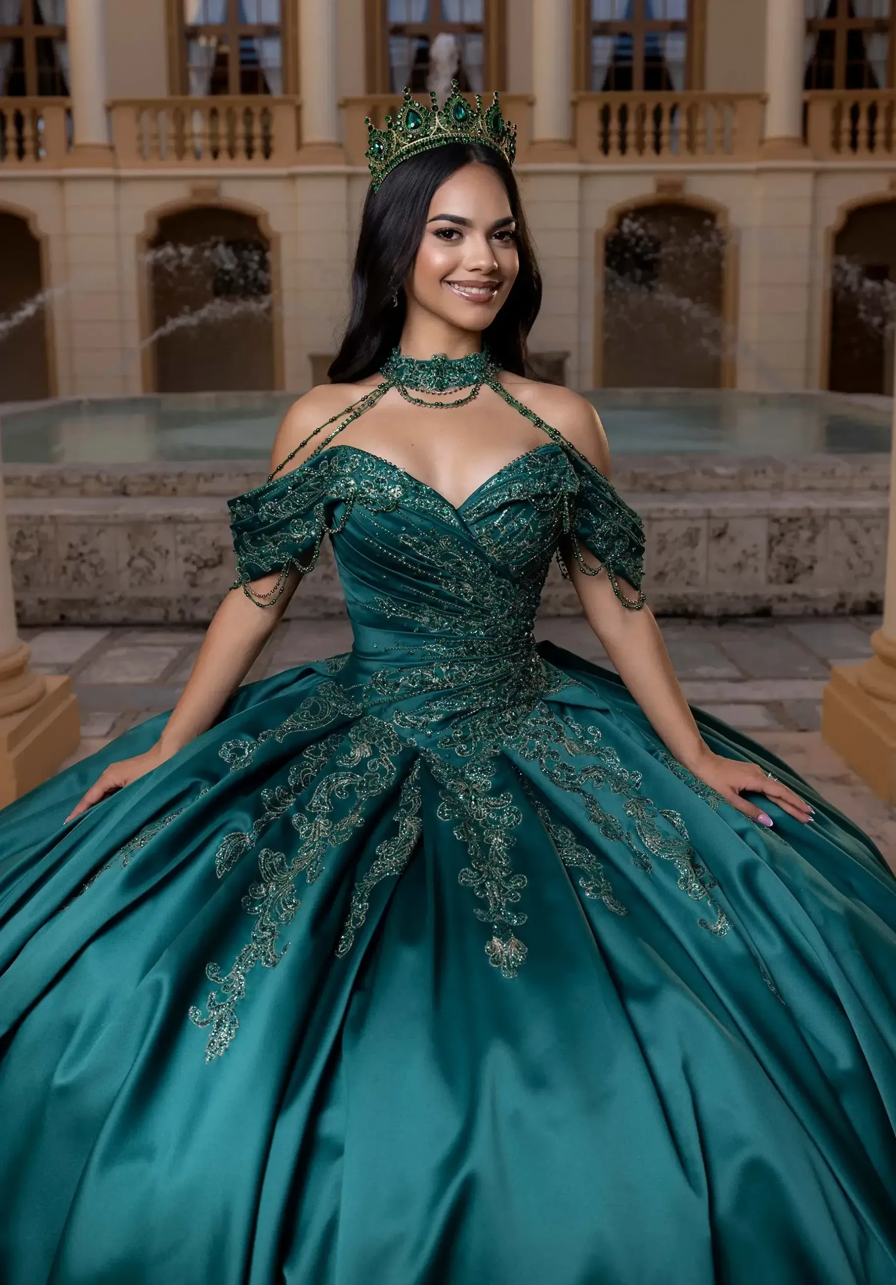 A woman in a detailed teal gown and crown stands in front of a fountain. The dress features intricate embroidery. She smiles confidently.