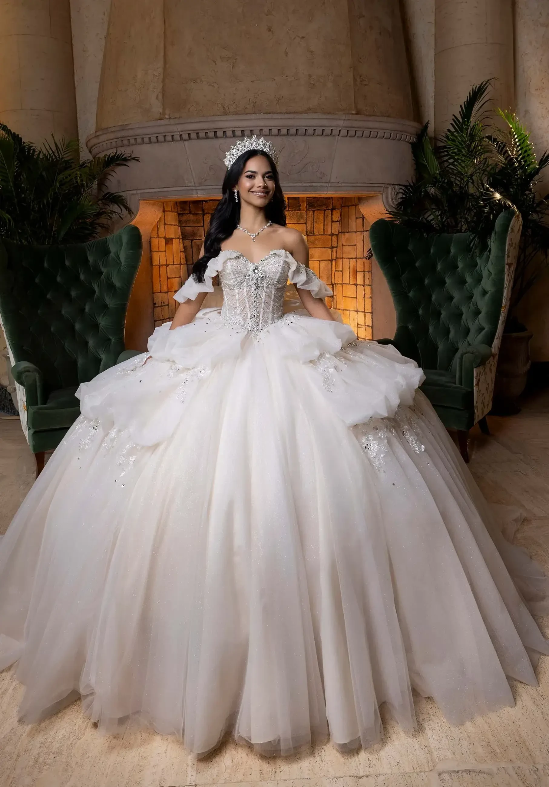 A young woman stands gracefully in a grand white ball gown with intricate details, wearing a tiara. She is in front of a large stone fireplace, flanked by green velvet chairs, exuding elegance and joy.