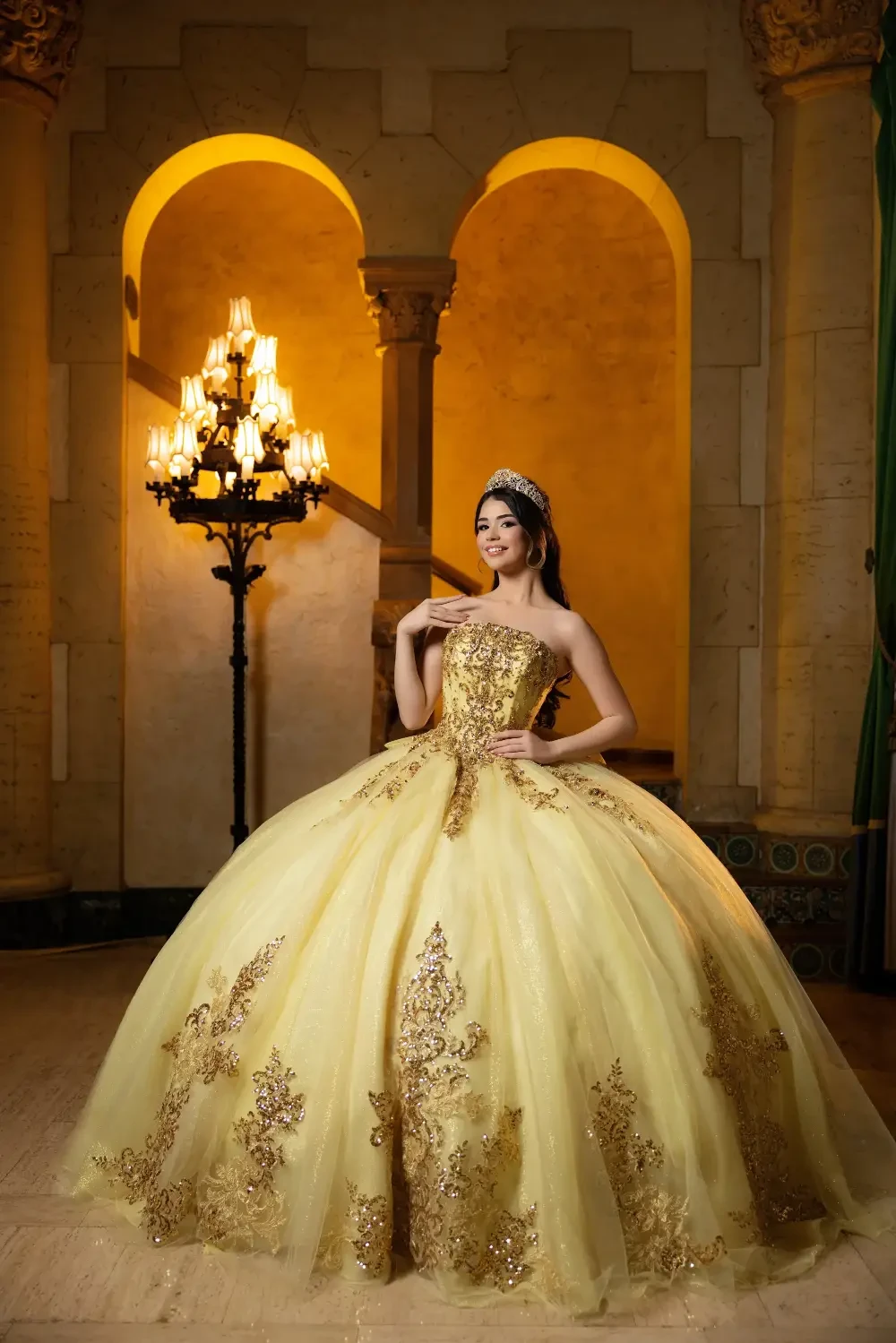 A person wearing a luxurious yellow ball gown with intricate gold detailing, standing gracefully in an elegant interior with chandeliers and an arched doorway.