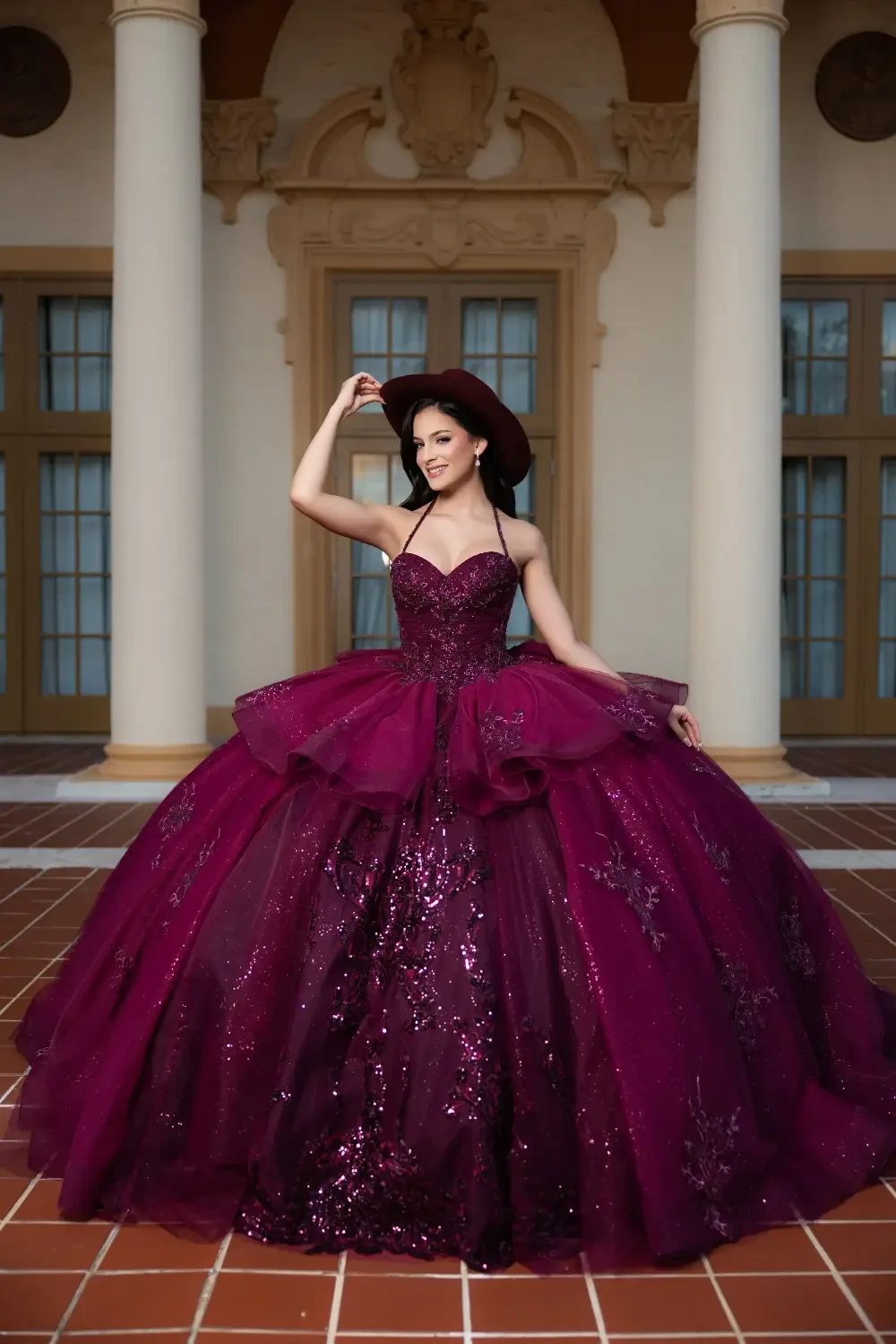 A woman in a burgundy ball gown with a fitted bodice and a full skirt stands gracefully, holding a hat and smiling in a grand architectural setting.