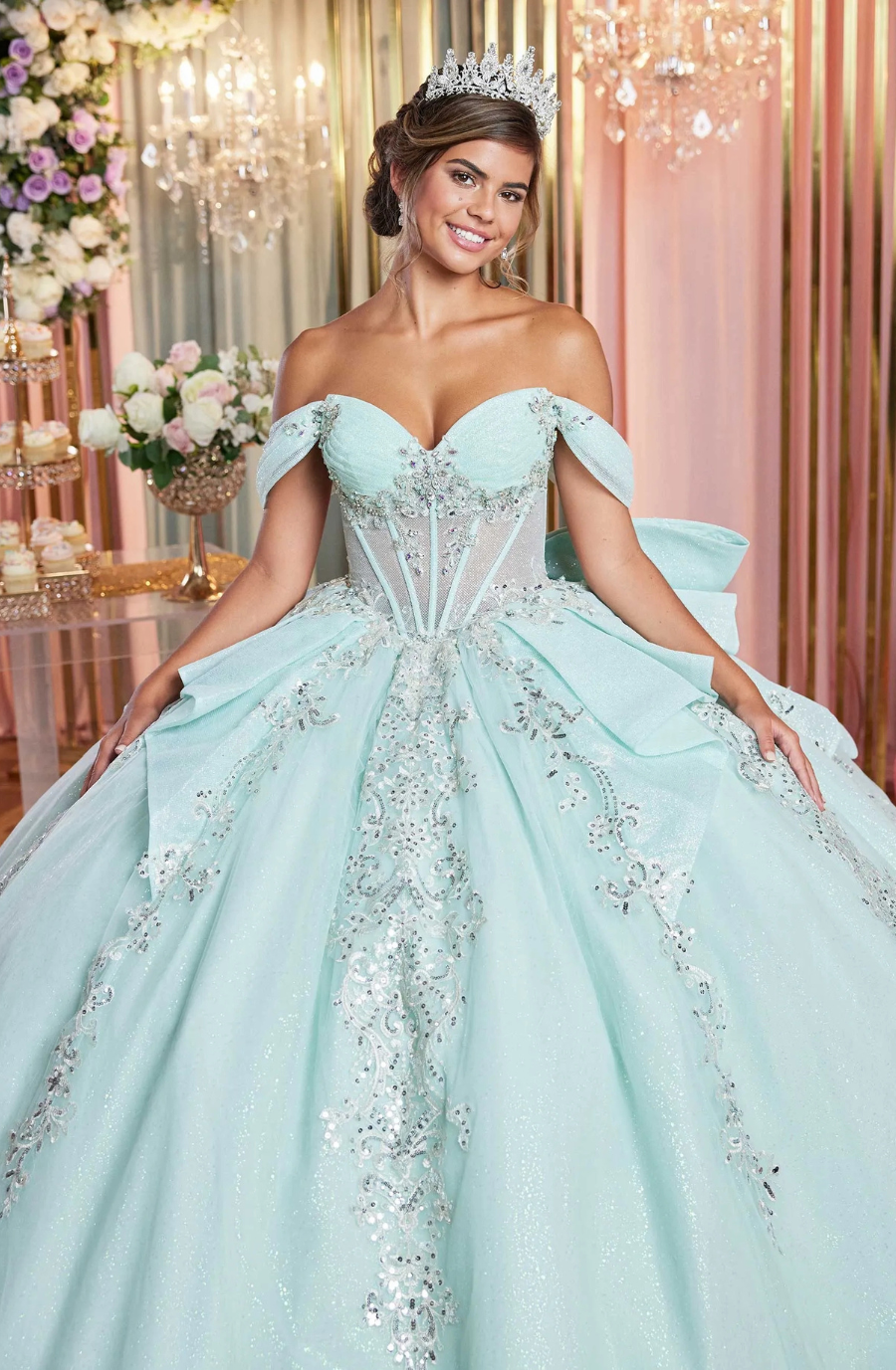 Model is wearing a mint green and silver ball gown with a strapless sweetheart neckline, sheer midriff with corset boning, glitter tulle skirt, waterfall peplum overlay, detachable off-the-shoulder sleeves, oversized bow, and sheer petal-shaped train.