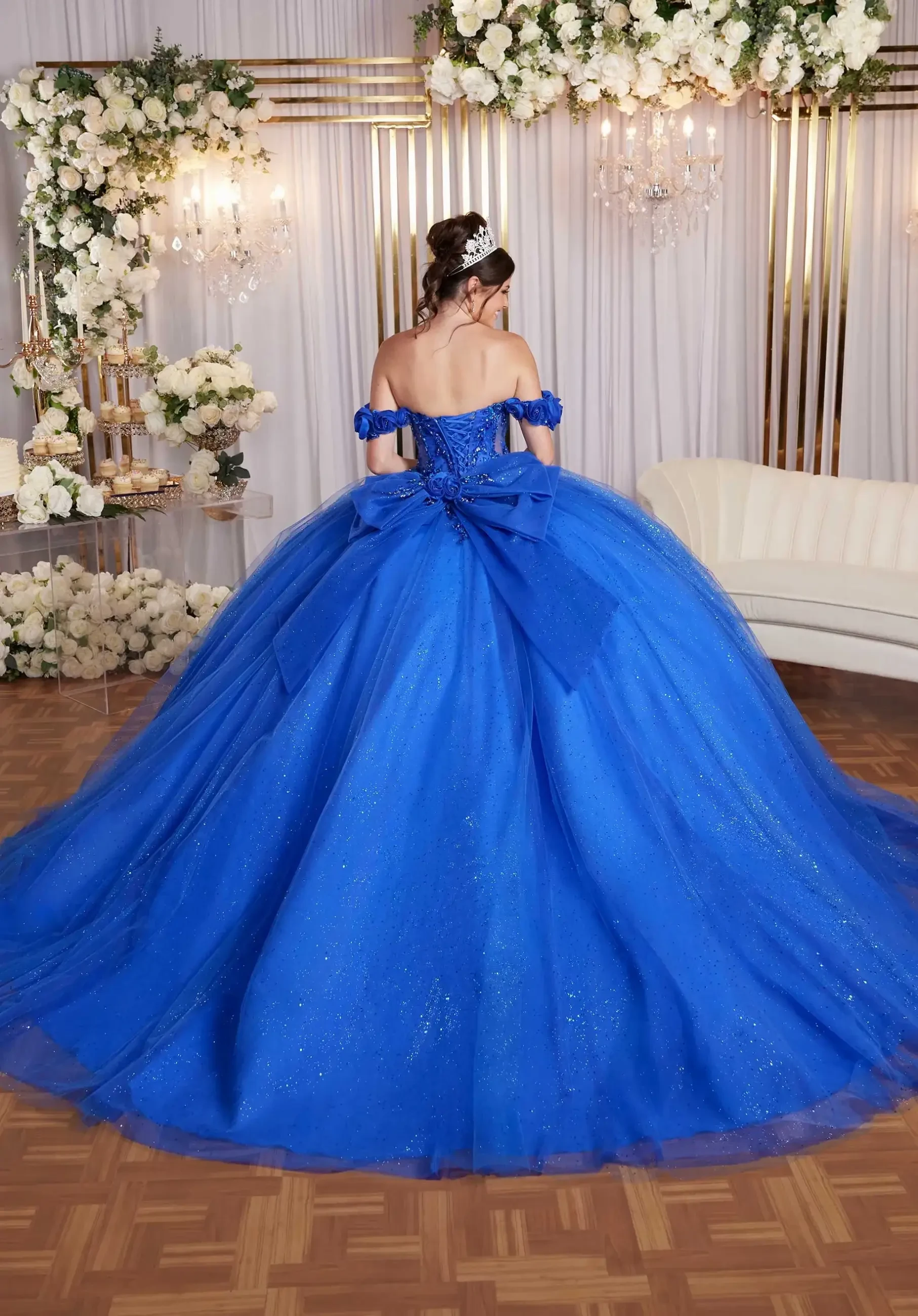 A woman in a sparkling blue ball gown with a large bow stands on a wooden floor. She faces floral decorations and chandeliers, exuding elegance and grace.