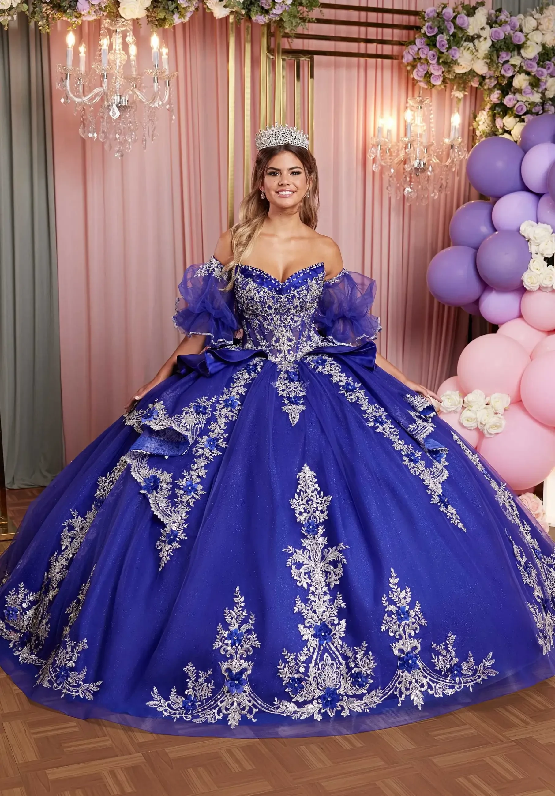 Model is wearing a royal purple ball gown with a cat-eye neckline, sheer boned bodice with crystal beading, basque waist, and a satin peplum skirt with cascading floral appliqués, detachable pouf sleeves, and an oversized satin bow.