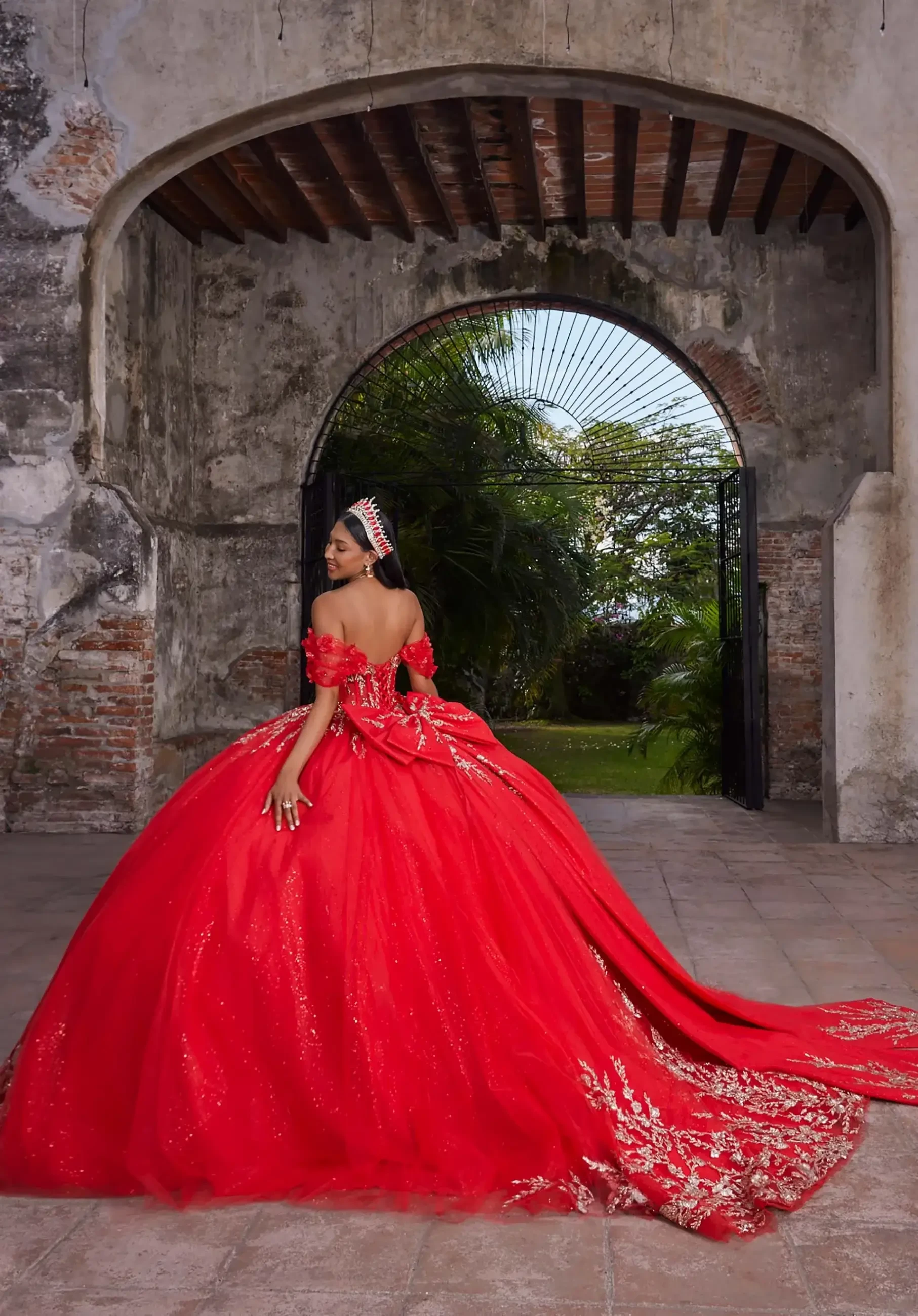 Woman in an elegant red ball gown with a tiara stands in a rustic archway, with lush greenery visible through the open gates. Regal and festive atmosphere.