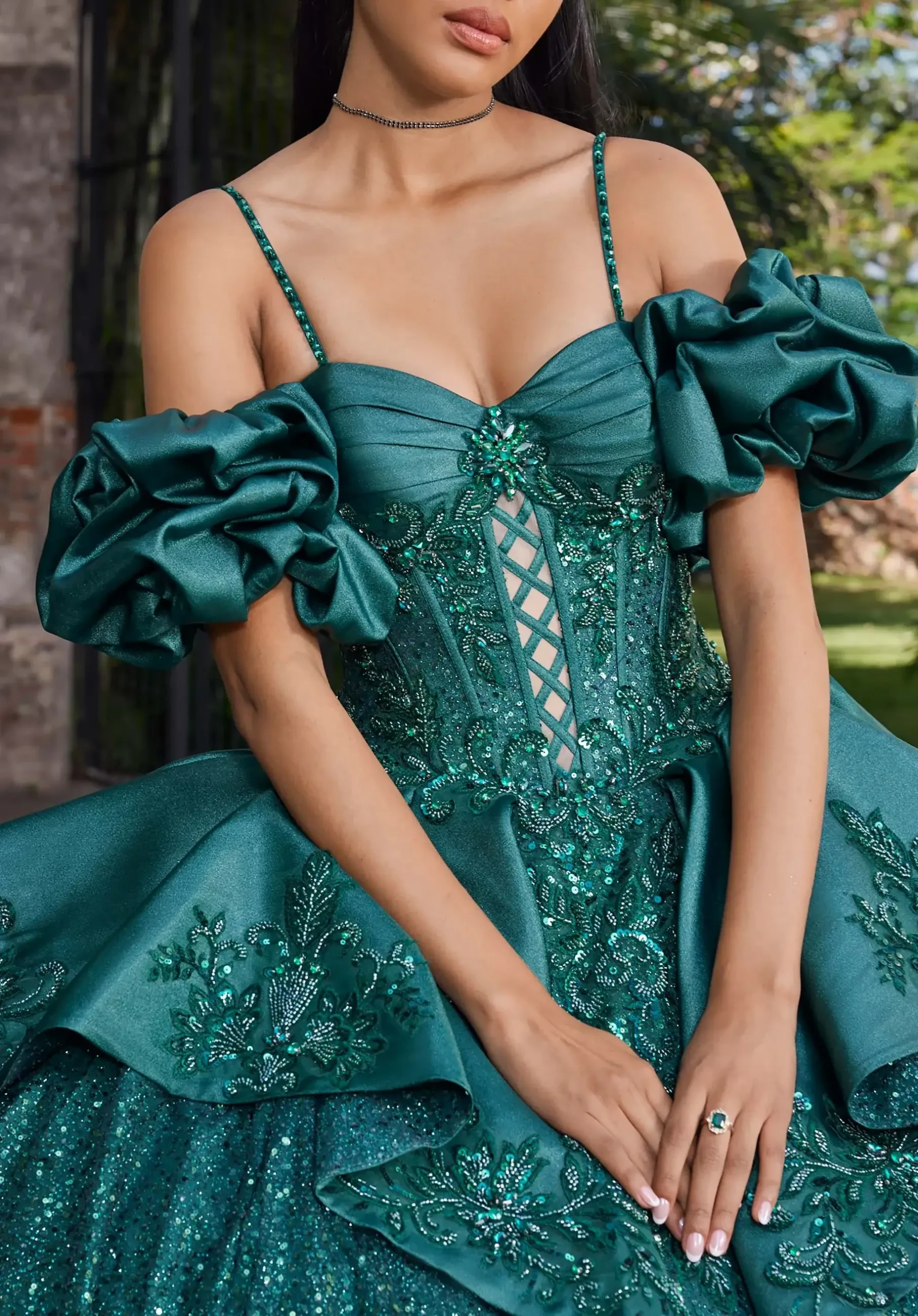 Woman in an ornate emerald-green gown with intricate beadwork and ruffled sleeves stands outdoors. The dress has a corset-style bodice and a sparkling skirt.