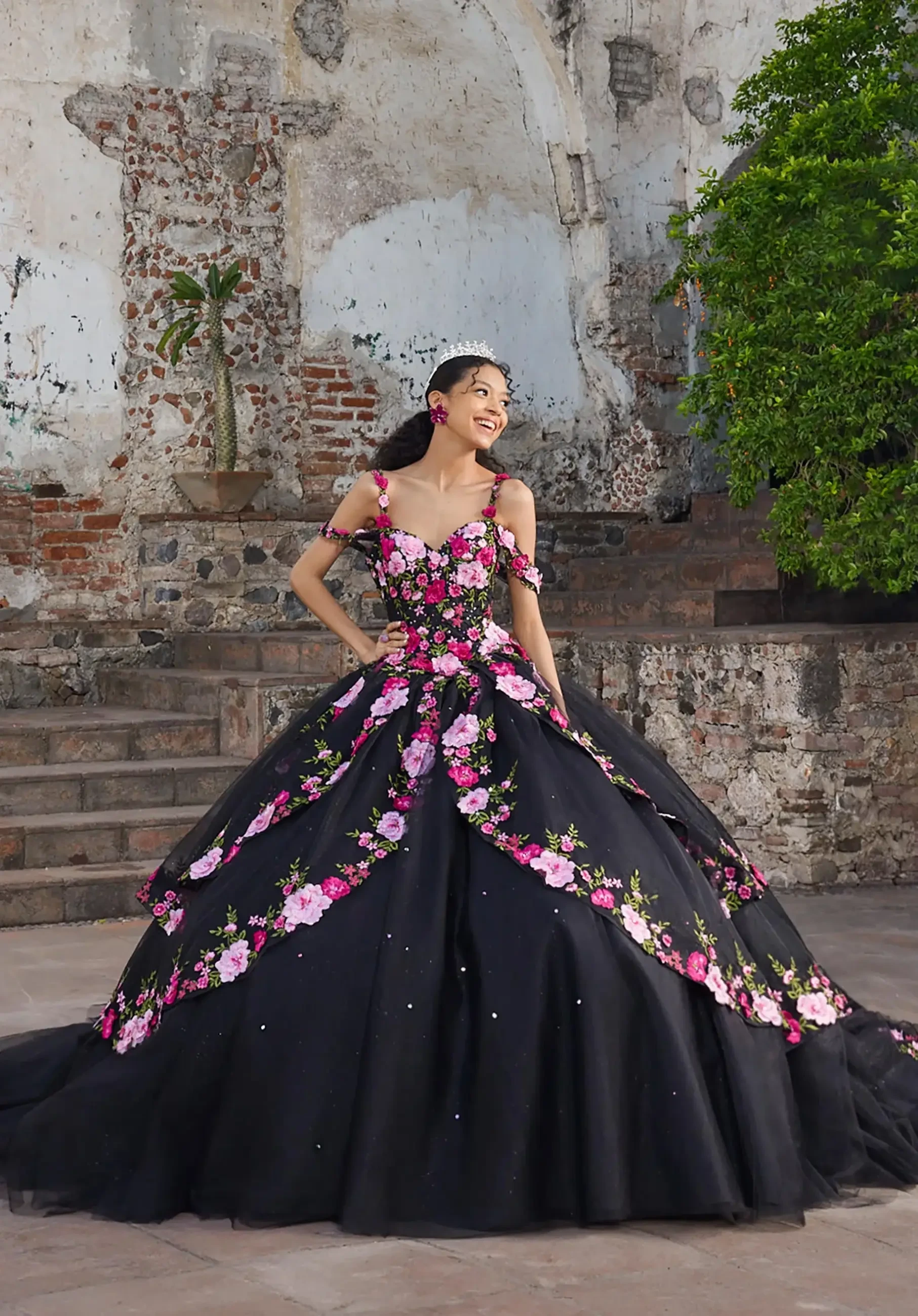 A woman smiles in a lavish black ball gown adorned with pink flowers. She stands on stone steps beside weathered brick walls and greenery. Elegant, joyful setting.