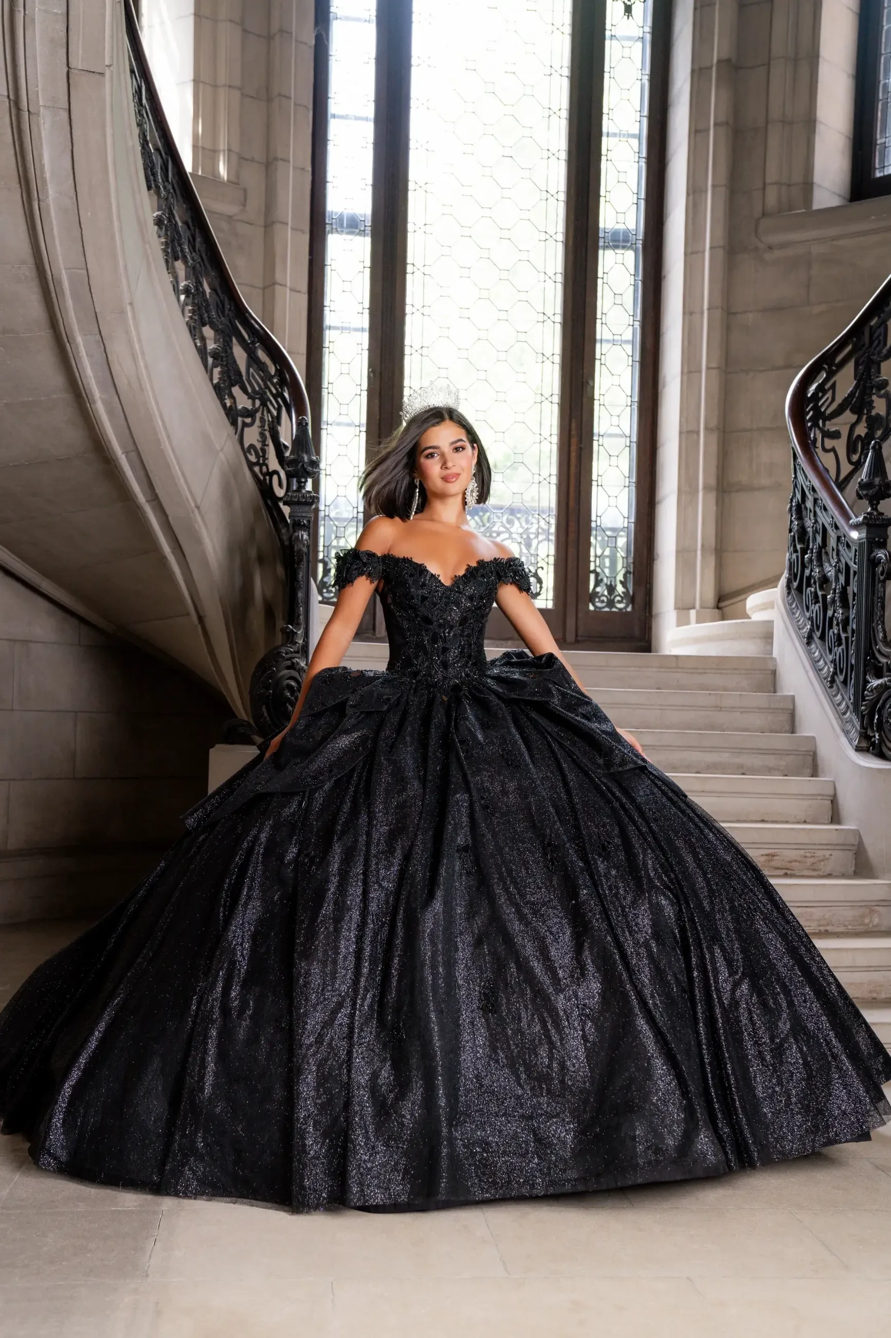 A woman in a shimmering black ball gown stands confidently on grand, ornate stairs. Sunlight streams through tall windows, adding elegance and grace.
