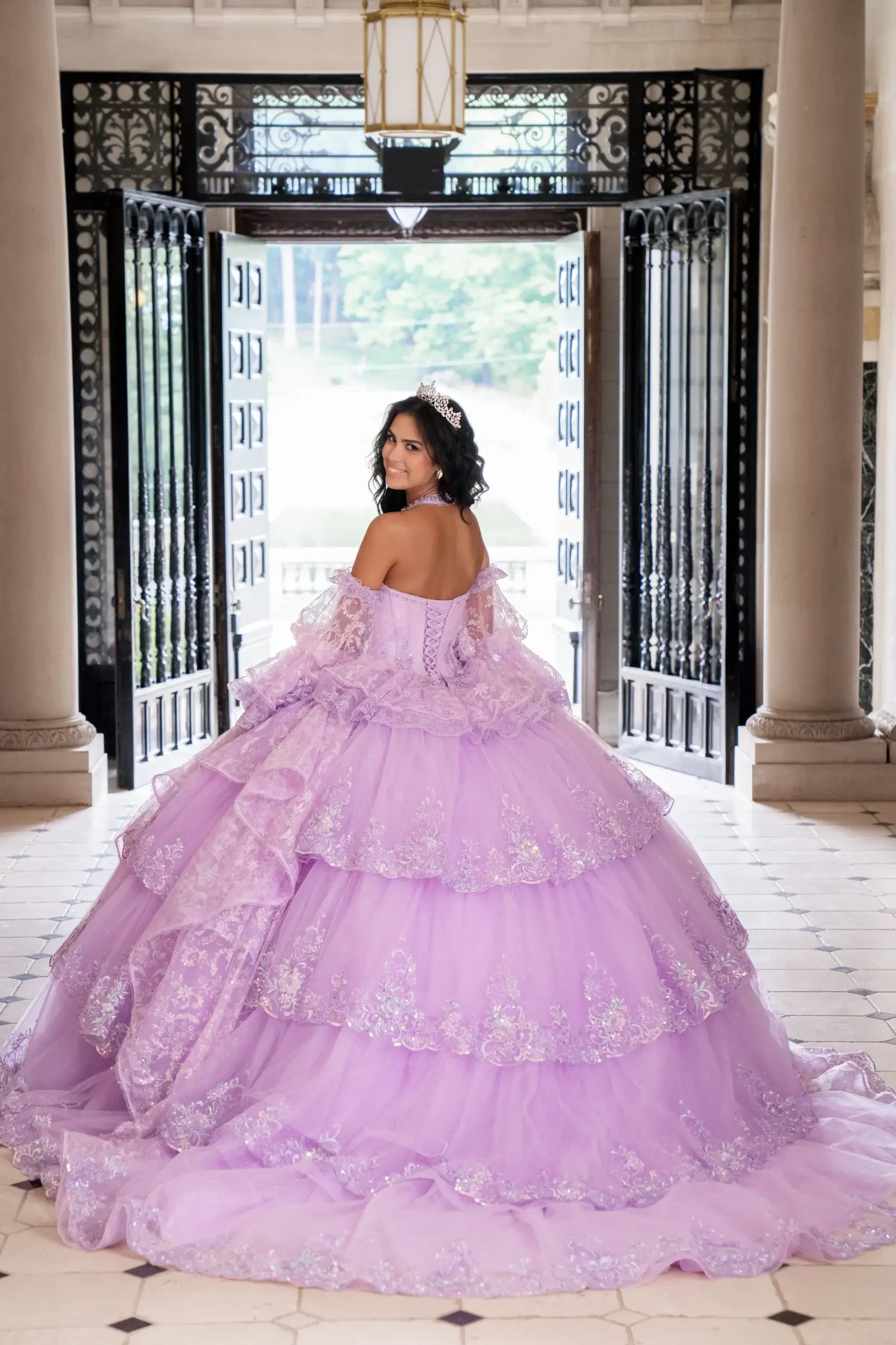 Model is wearing a lilac ball gown with tiered tulle layers, sequined appliqués, a sweetheart bodice, and detachable sleeves, styled with a matching necklace.