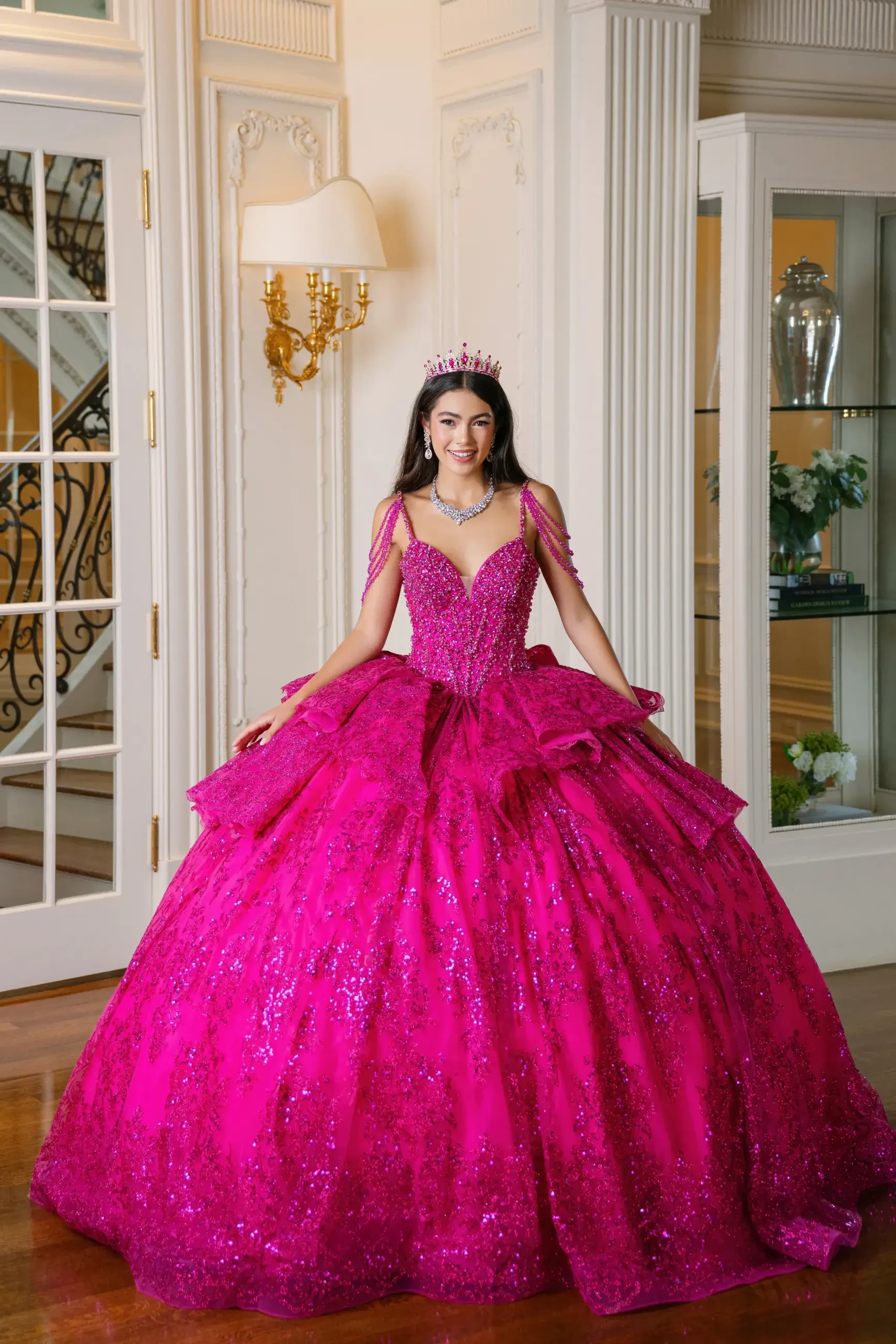 A person wearing a large, bright pink ball gown with intricate embellishments and a tiara, standing in an elegant room with light-colored walls and decorative elements.