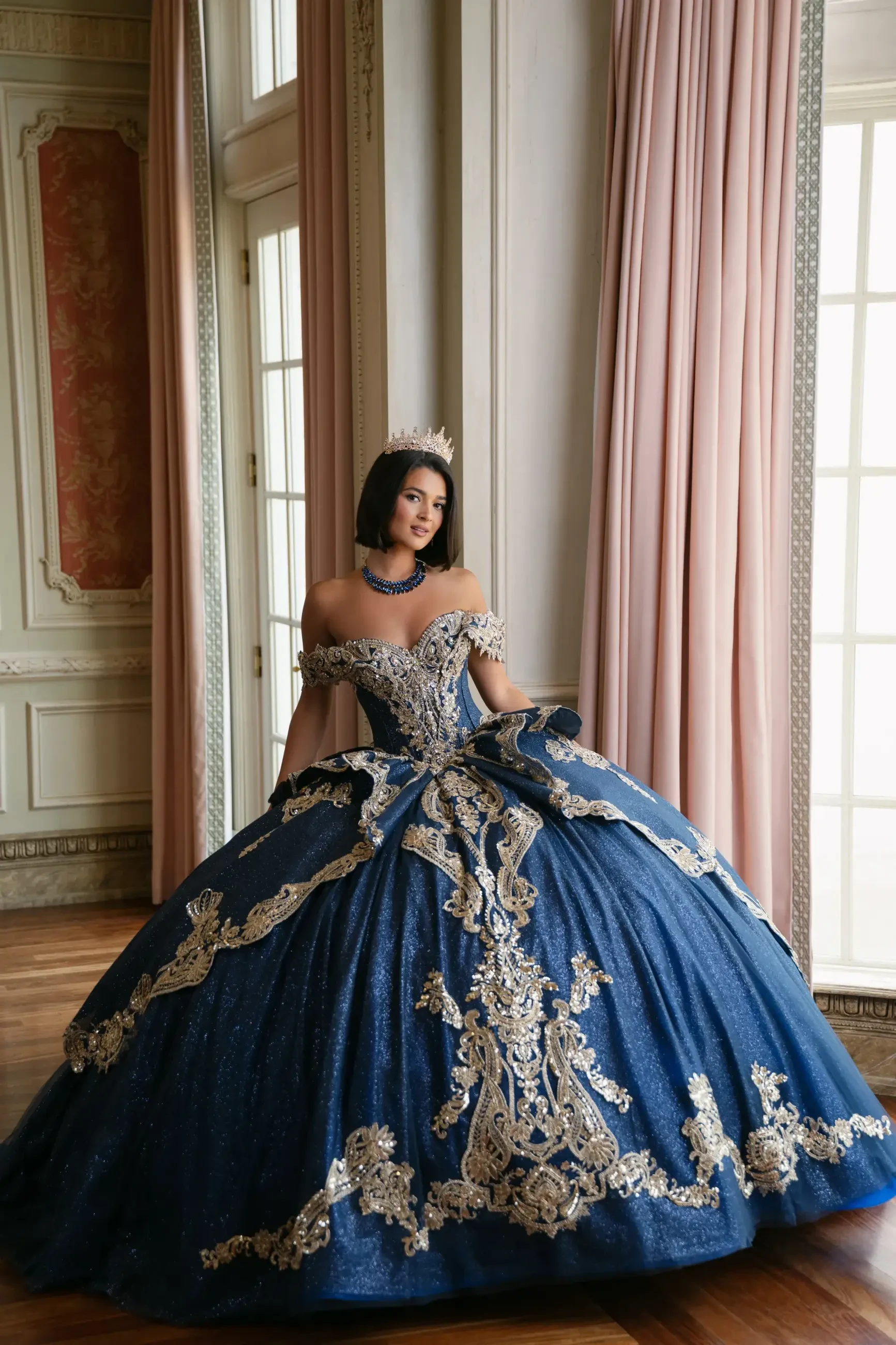 A woman wearing a lavish blue gown with ornate gold embroidery, standing gracefully in a well-lit room with elegant curtains and vintage decor.