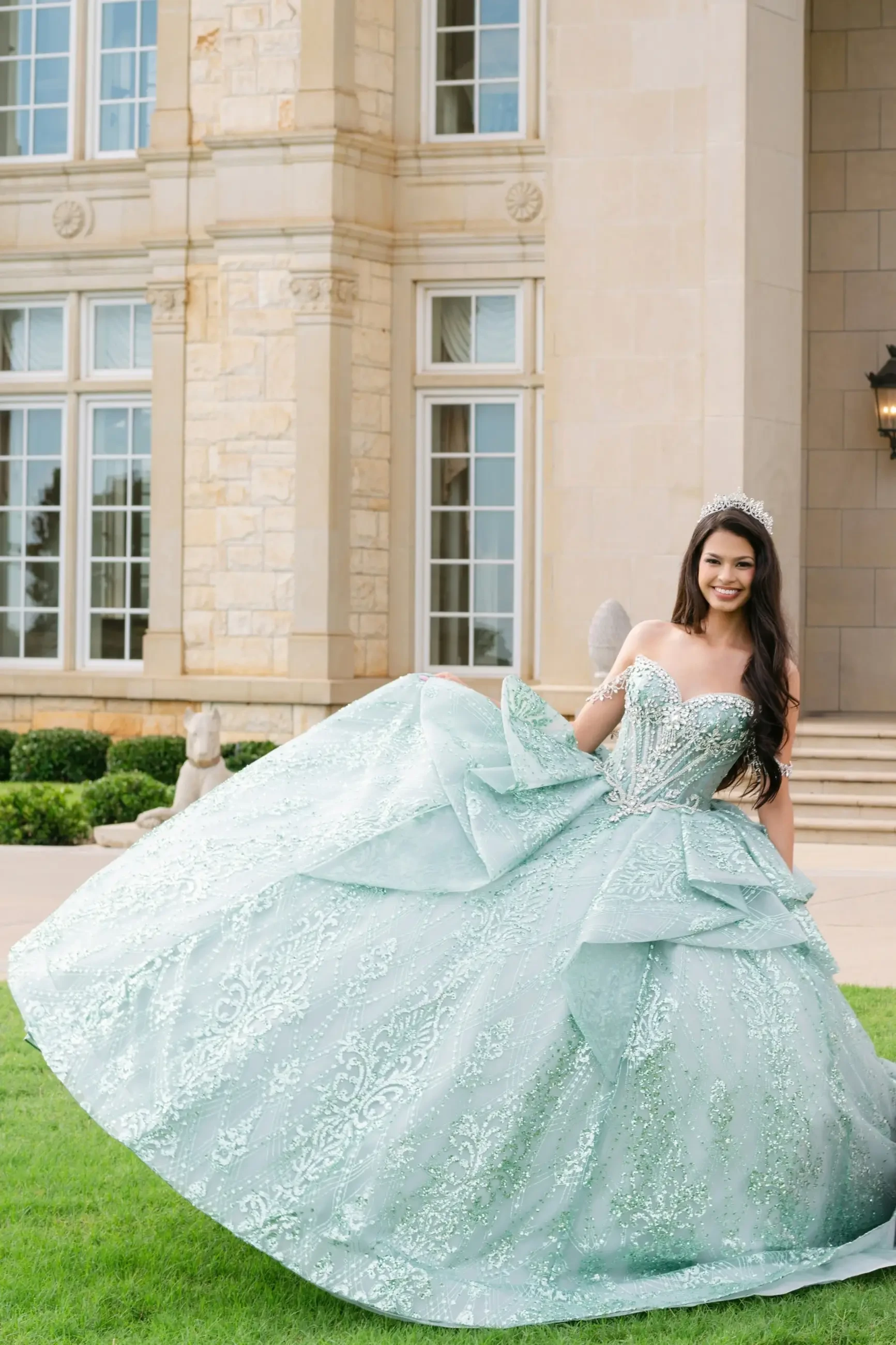 A woman in a stunning mint green ball gown stands outside a large, elegant building. The dress features intricate designs and a full skirt, and she is smiling while holding the skirt of her dress.