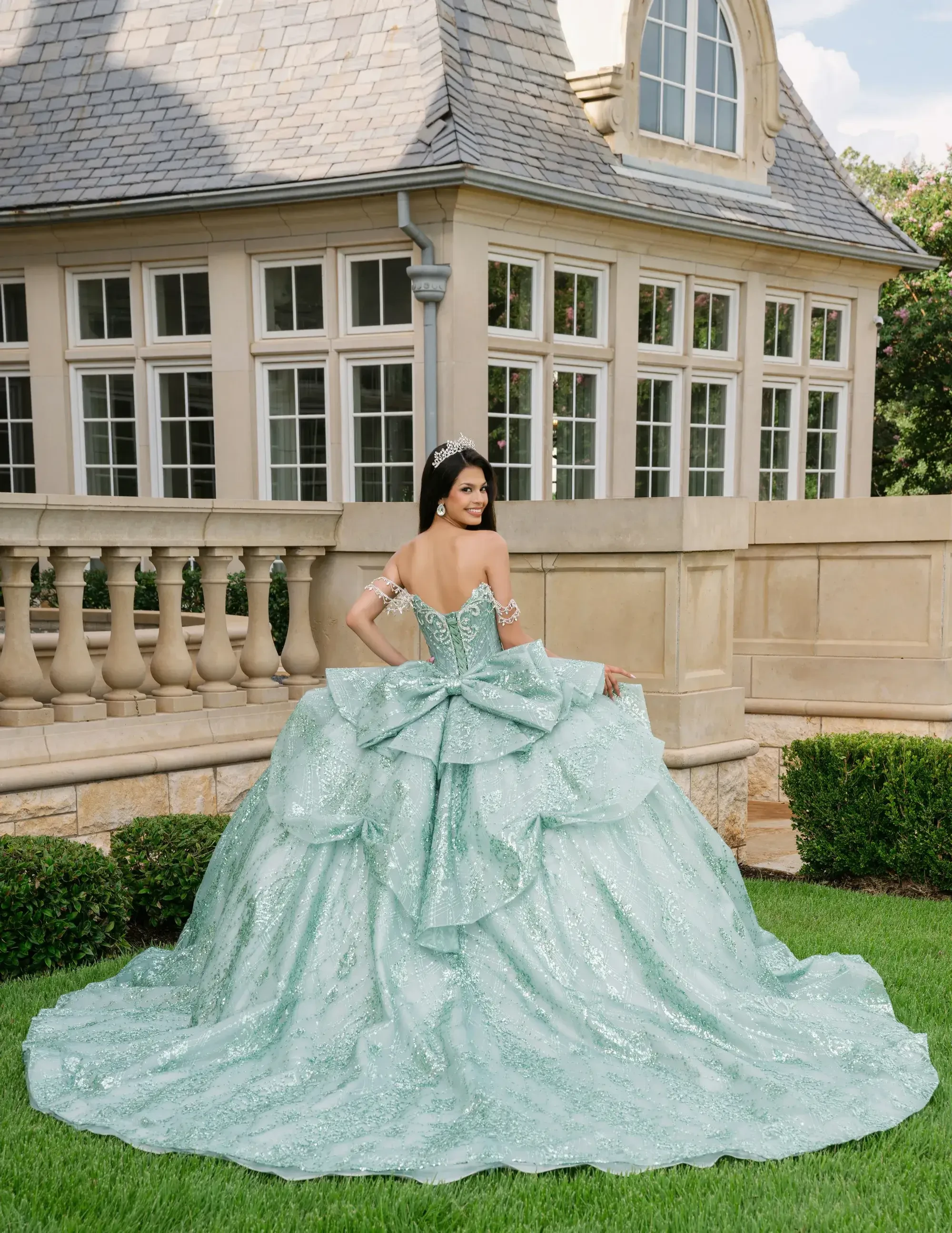 A person wearing a large, sparkling mint green gown with a big bow on the back, standing in a grassy area near an elegant building with large windows.